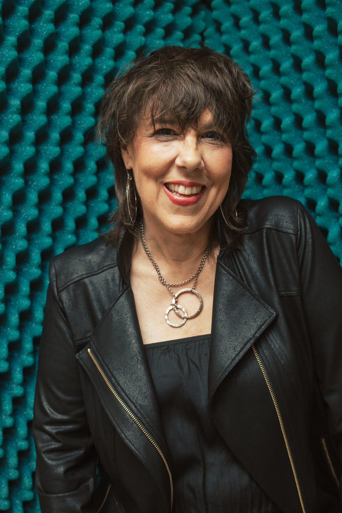 Beth Falcone, smiling, in a black leather jacket in front of acoustic foam in a recording studio.