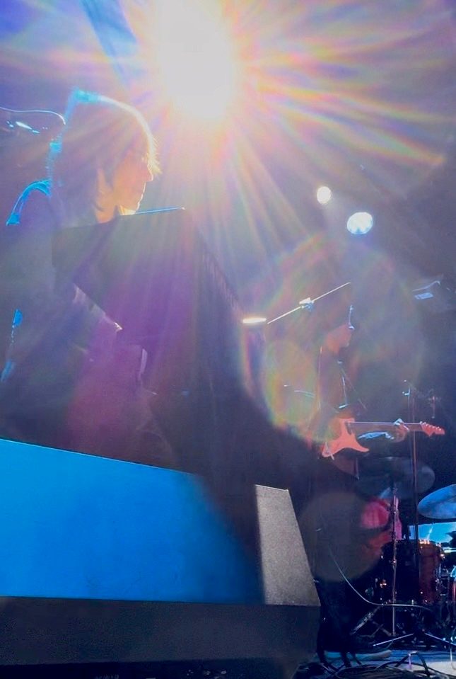 Beth at the keyboard on stage, backlit by a stage light, with the band behind her.