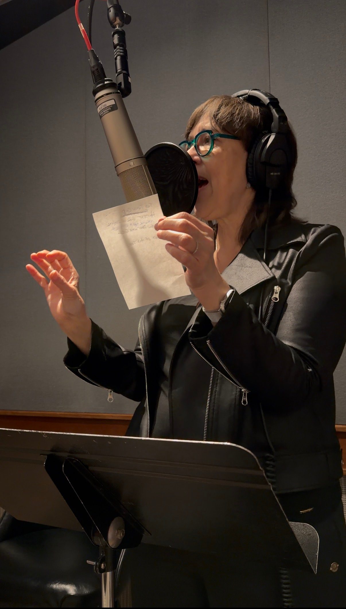 Beth singing into a studio microphone, holding a lyric sheet.