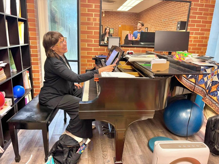 Beth laughing at a grand piano in a teaching studio, with students reflected in the mirror.