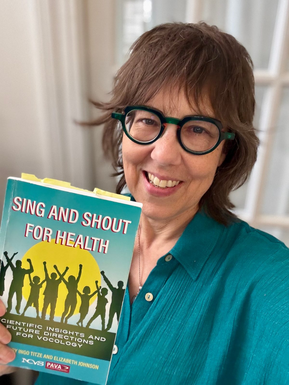 Beth smiling and holding a copy of Sing and Shout for Health: Scientific Insights and Future Directions for Vocology.